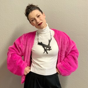 May include: A woman wearing a hot pink faux fur jacket over a white ribbed turtleneck and black pants. A black geometric pendant necklace hangs from a black cord. The background is a neutral gray wall.