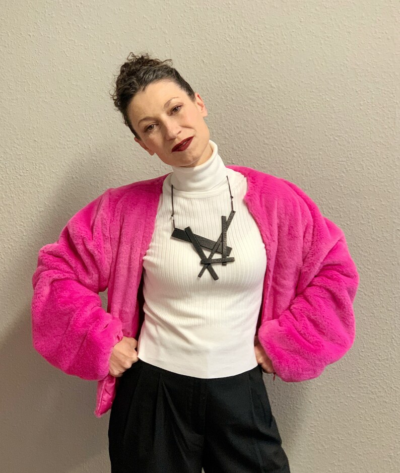 May include: A woman wearing a hot pink faux fur jacket over a white ribbed turtleneck and black pants. A black geometric pendant necklace hangs from a black cord. The background is a neutral gray wall.