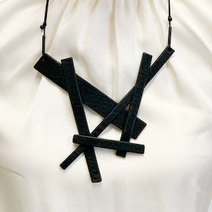 May include: A black geometric necklace featuring overlapping rectangular shapes. The pendant hangs from a thin, dark cord. The necklace is displayed against a white background, highlighting the contrast of the dark accessory.