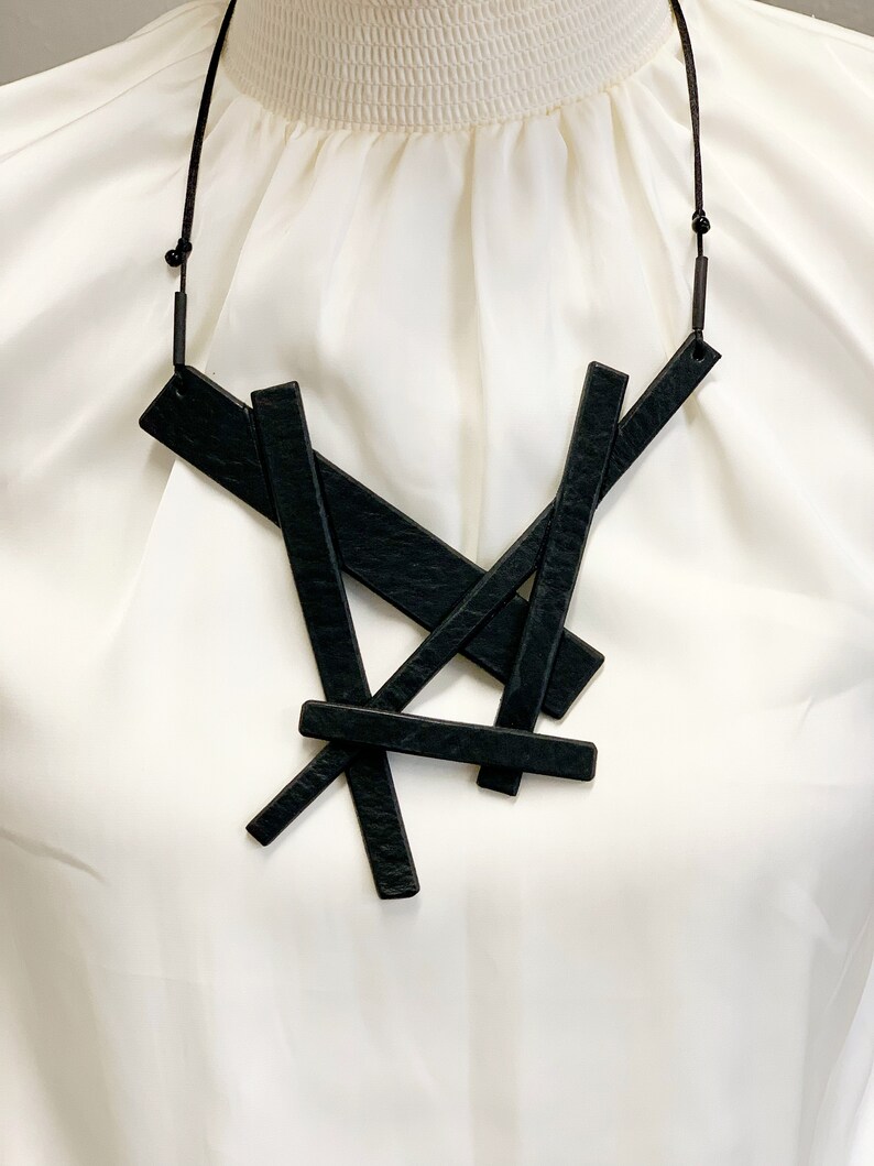 May include: A black geometric necklace featuring overlapping rectangular shapes. The pendant hangs from a thin, dark cord. The necklace is displayed against a white background, highlighting the contrast of the dark accessory.