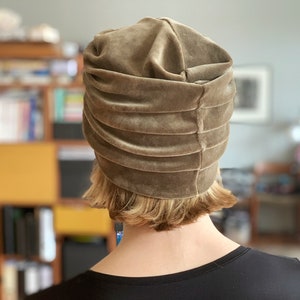 May include: A taupe-colored velvet turban hat with horizontal pleats. The hat is worn by a person with short, light-colored hair and a black top. The background is blurred, showing a room with bookshelves and artwork.