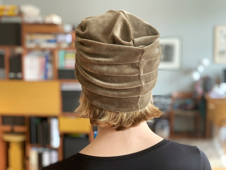 May include: A taupe-colored velvet turban hat with horizontal pleats. The hat is worn by a person with short, light-colored hair and a black top. The background is blurred, showing a room with bookshelves and artwork.