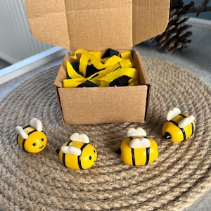 Puede incluir: Cuatro pequeñas figuras de abejorros amarillos y negros con alas blancas dispuestas frente a una caja de cartón marrón abierta llena de relleno de papel amarillo y negro. Cada abeja tiene un cuerpo redondeado, rayas negras y una cara sonriente.