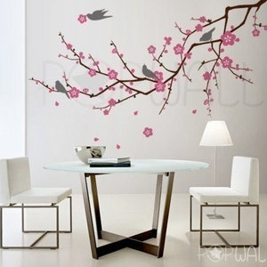 May include: A white wall with a pink and gray cherry blossom branch decal. The decal features small birds and pink flowers. Two white chairs and a glass table are in front of the wall.