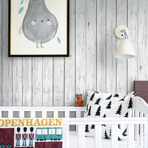 May include: White wood panel wallpaper with a framed illustration of a grey pear with a smiling face. A white cot with a white and black patterned pillow and a white blanket with the text "COPENHAGEN" and a colourful illustration of buildings and a bus. The text "NOWALL PEEL & STICK REMOVABLE WALLPAPER" is visible in the bottom right corner.