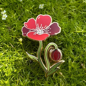 May include: A red and burgundy enamel poppy flower pin with gold-colored trim. The pin features a detailed design of a blooming flower and a bud, set against a backdrop of green moss.