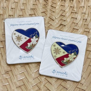 May include: Two heart-shaped enamel pins featuring the Filipino flag design. The pins are coloured blue, yellow, and red, with a gold sun and stars. The cards are printed with the text "PUSONG PINOY" and "filipino heart enamel pin".
