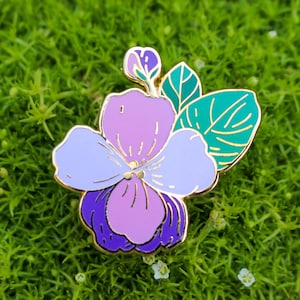 May include: A floral enamel pin featuring a violet flower with purple, lavender, and white petals, accented with gold trim. Two green leaves with gold outlines are on the right. The pin is set against a green background.