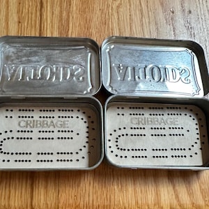 Può includere: Scatole di metallo aperte con la scritta "SCOTTS" in rilievo sul coperchio. Il fondo delle scatole contiene una carta con la scritta "CRIBBAGE" e un motivo di fori per segnare il gioco. Le scatole sono aperte e visualizzate su una superficie di legno.