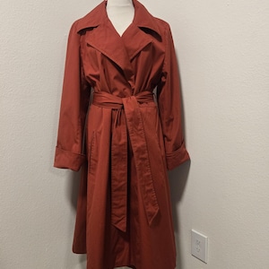 May include: A long, rust-colored trench coat featuring a wide notched collar, long sleeves with folded cuffs, and a matching fabric tie belt at the waist. The coat has a structured silhouette and falls to a midi length, displayed on a mannequin.