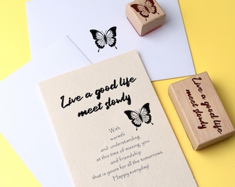 May include: Two wooden stamps with the text "Live a good life meet slowly" and a butterfly design. The stamps are on a white card with a handwritten message.