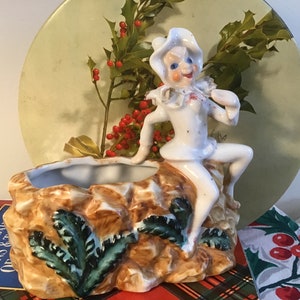 May include: A vintage ceramic planter featuring a seated figure in white attire with a hat, perched on a rock formation. The planter has a trough for plants and is decorated with green fern-like accents. The figure has a smiling face.