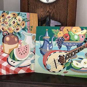 May include: Two vintage paint-by-number canvases. One features a still life with sunflowers, watermelon, and a pitcher on a red and white checkered tablecloth. The other depicts a mandolin, fruit bowl, and a blue teapot. The paintings have a retro aesthetic.
