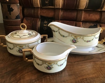 May include: A three-piece porcelain serving set with a sugar bowl, creamer, and gravy boat. The set features a pale yellow background with floral garland and gold trim. The sugar bowl has a lid with a gold handle.