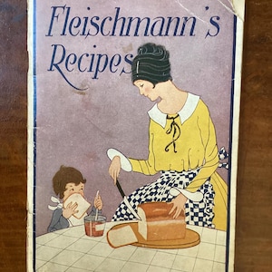 May include: A vintage Fleischmann's Recipes booklet featuring a color illustration of a person in a yellow shirt and blue and white checkered apron slicing a loaf of bread on a wooden board, with a child eating bread at a table in the foreground.
