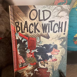 May include: A vintage children's book titled "Old Black Witch!" features a cartoon illustration of a witch flying on a broom. The cover art shows the witch exiting a broken window. The book is propped up on a small black stand.