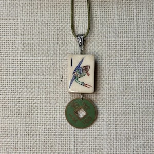 May include: A distinctive pendant necklace. It features a rectangular tile with a colourful bird design and the number 1. Below the tile hangs a green coin with a square centre. The necklace is strung on a green cord.