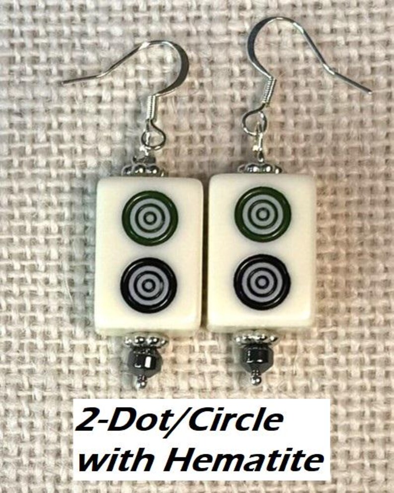 May include: Earrings with rectangular white tiles, each displaying green and black concentric circles. The earrings have silver hooks and hematite bead details. The text "2-Dot/Circle with Hematite" is visible below the earrings.
