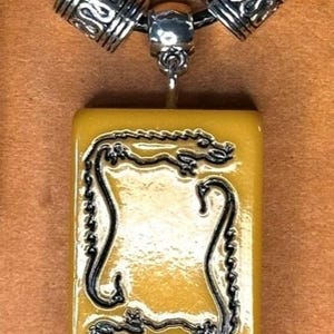 Puede incluir: Un collar con colgante que presenta un dije rectangular de color amarillo con un dise&ntilde;o de drag&oacute;n negro. El dije cuelga de un cord&oacute;n negro con cuentas decorativas plateadas. El texto "White Dragon" se muestra en la parte inferior.