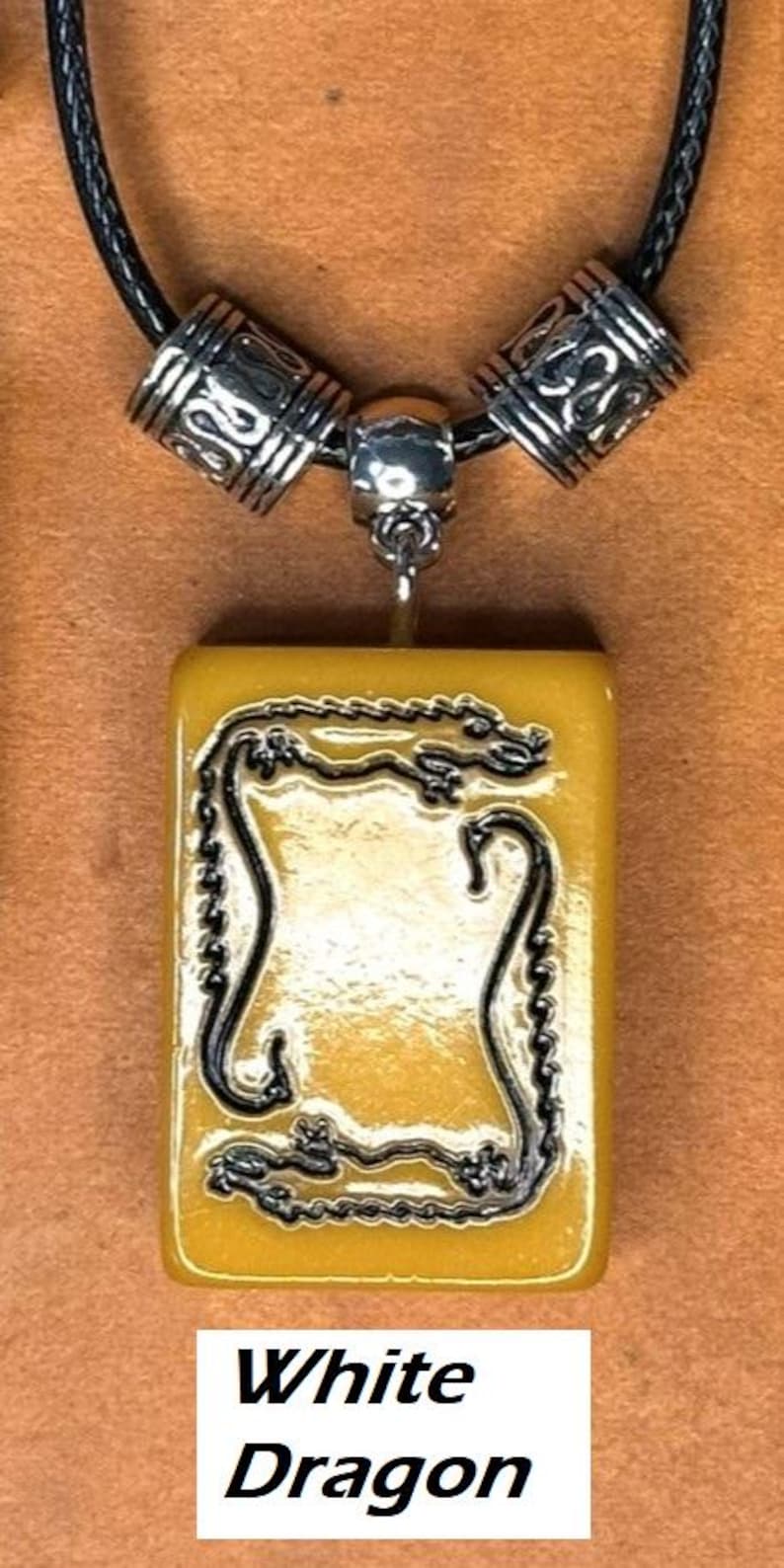 Puede incluir: Un collar con colgante que presenta un dije rectangular de color amarillo con un dise&ntilde;o de drag&oacute;n negro. El dije cuelga de un cord&oacute;n negro con cuentas decorativas plateadas. El texto "White Dragon" se muestra en la parte inferior.
