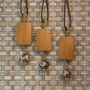 Puede incluir: Tres collares con colgantes rectangulares de madera y colgantes decorativos de esmalte con forma de linterna de colores. Los colgantes est&aacute;n sujetos a cordones marrones con cierres dorados. El fondo es un patr&oacute;n tejido en tonos marrones y beige.