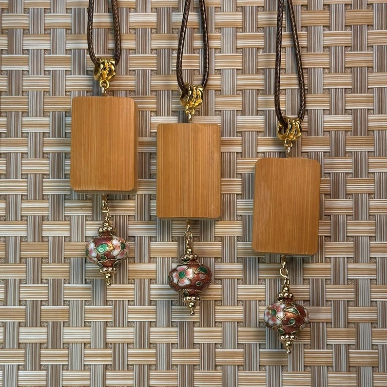 Puede incluir: Tres collares con colgantes rectangulares de madera y colgantes decorativos de esmalte con forma de linterna de colores. Los colgantes est&aacute;n sujetos a cordones marrones con cierres dorados. El fondo es un patr&oacute;n tejido en tonos marrones y beige.