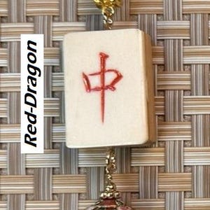 Puede incluir: Un collar con una ficha de Mahjong crema con un car&aacute;cter rojo y un colgante decorativo. El colgante dorado presenta dise&ntilde;os florales rojos, verdes y blancos. El collar tiene un cord&oacute;n marr&oacute;n.