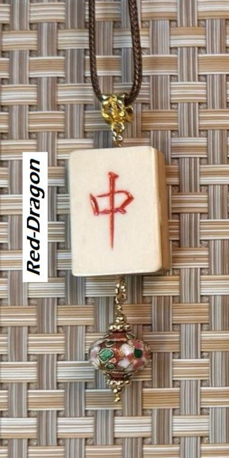 Puede incluir: Un collar con una ficha de Mahjong crema con un car&aacute;cter rojo y un colgante decorativo. El colgante dorado presenta dise&ntilde;os florales rojos, verdes y blancos. El collar tiene un cord&oacute;n marr&oacute;n.