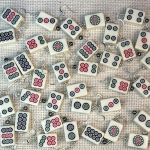 May include: A collection of ivory-colored Mahjong tile earrings with silver-tone hooks. Each earring features a different design with red, green, and black details. The earrings are scattered across a textured, off-white surface.