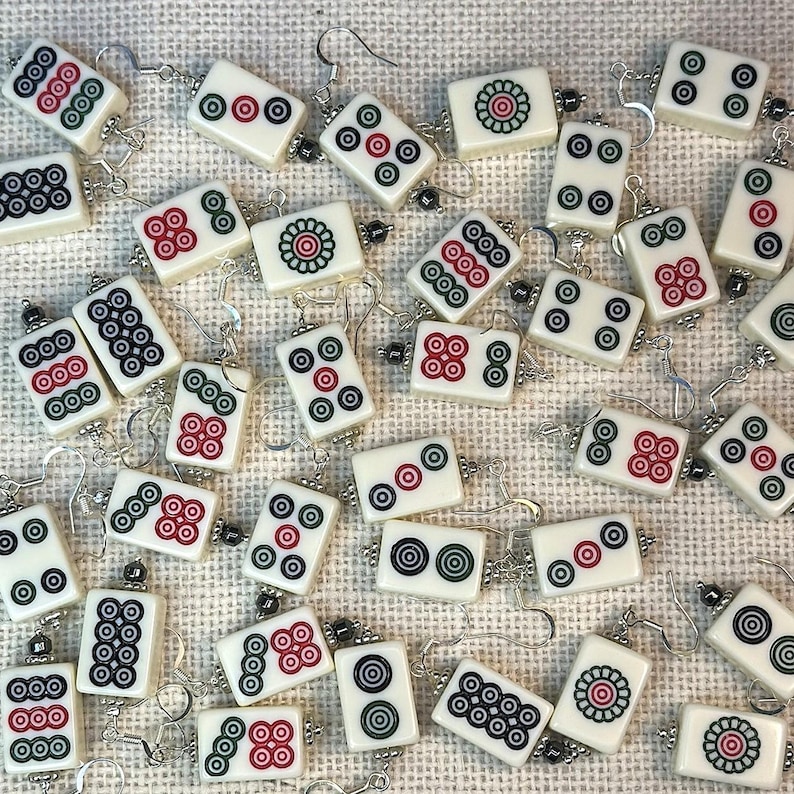 May include: Ivory-coloured Mahjong tile earrings with silver-tone hooks. Each earring displays a unique design with red, green, and black details. The earrings are scattered across a textured, off-white surface.