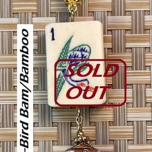 Puede incluir: Un collar con colgante que presenta una ficha de Mahjong vintage con un dise&ntilde;o de p&aacute;jaro y bamb&uacute;. La ficha es de color crema con detalles azules y verdes. El colgante est&aacute; conectado a un cord&oacute;n marr&oacute;n y tiene un cierre decorativo dorado y un dije con estampado floral.