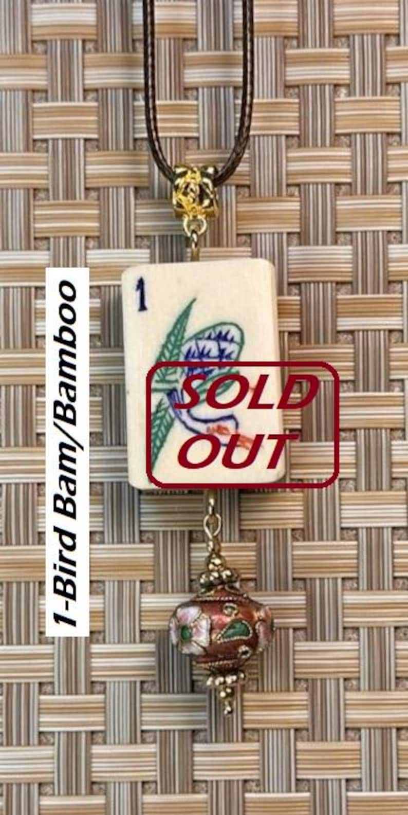 Puede incluir: Un collar con colgante que presenta una ficha de Mahjong vintage con un dise&ntilde;o de p&aacute;jaro y bamb&uacute;. La ficha es de color crema con detalles azules y verdes. El colgante est&aacute; conectado a un cord&oacute;n marr&oacute;n y tiene un cierre decorativo dorado y un dije con estampado floral.