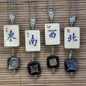Op de afbeelding: Vier zilveren kettingkettingen met hangers. Elke hanger heeft een rechthoekige tegel met Chinese karakters en een vierkante, veelzijdige kraal. De tegels zijn crèmekleurig met blauwe karakters. De kralen zijn donkerrood en zilver.