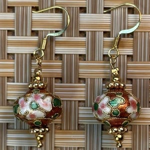 Puede incluir: Un par de pendientes dorados con colgantes en forma de linterna. Los colgantes est&aacute;n decorados con esmalte marr&oacute;n, blanco, rosa y verde, con detalles dorados. Los pendientes se muestran sobre un fondo tejido.