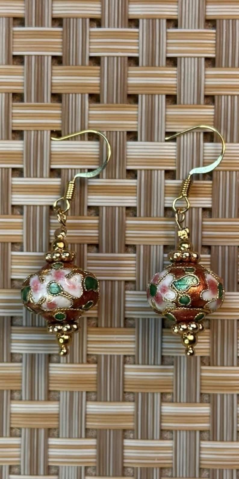 Puede incluir: Un par de pendientes dorados con colgantes en forma de linterna. Los colgantes est&aacute;n decorados con esmalte marr&oacute;n, blanco, rosa y verde, con detalles dorados. Los pendientes se muestran sobre un fondo tejido.