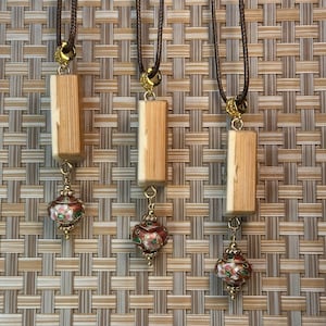 Puede incluir: Tres collares con cordones marrones, detalles dorados y colgantes rectangulares de madera. Cada colgante se combina con un peque&ntilde;o dije en forma de linterna esmaltada roja y verde. El fondo es un patr&oacute;n tejido.