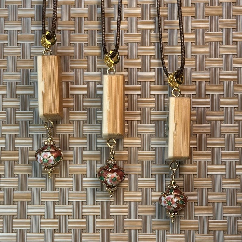 Puede incluir: Tres collares con cordones marrones, detalles dorados y colgantes rectangulares de madera. Cada colgante se combina con un peque&ntilde;o dije en forma de linterna esmaltada roja y verde. El fondo es un patr&oacute;n tejido.