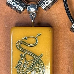 Puede incluir: Un collar con colgante que presenta un colgante rectangular de color amarillo dorado con un dise&ntilde;o de drag&oacute;n verde. El colgante est&aacute; suspendido de un collar de cord&oacute;n negro con cuentas decorativas plateadas. El texto "Green Dragon" se muestra en la parte inferior de la imagen.