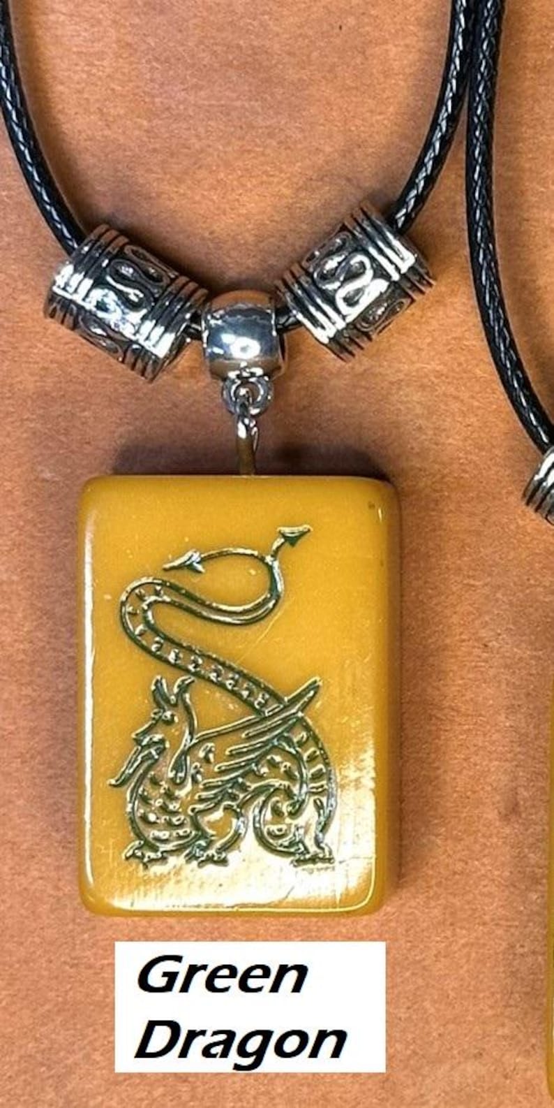 Puede incluir: Un collar con colgante que presenta un colgante rectangular de color amarillo dorado con un dise&ntilde;o de drag&oacute;n verde. El colgante est&aacute; suspendido de un collar de cord&oacute;n negro con cuentas decorativas plateadas. El texto "Green Dragon" se muestra en la parte inferior de la imagen.