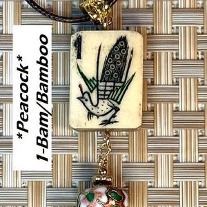 Puede incluir: Un collar con colgante que presenta una baldosa de color crema con una ilustraci&oacute;n de pavo real y el texto "1-Bam/Bamboo". El colgante est&aacute; conectado a una cuenta decorativa esmaltada y una cadena de metal dorado. El collar cuelga de un cord&oacute;n negro.