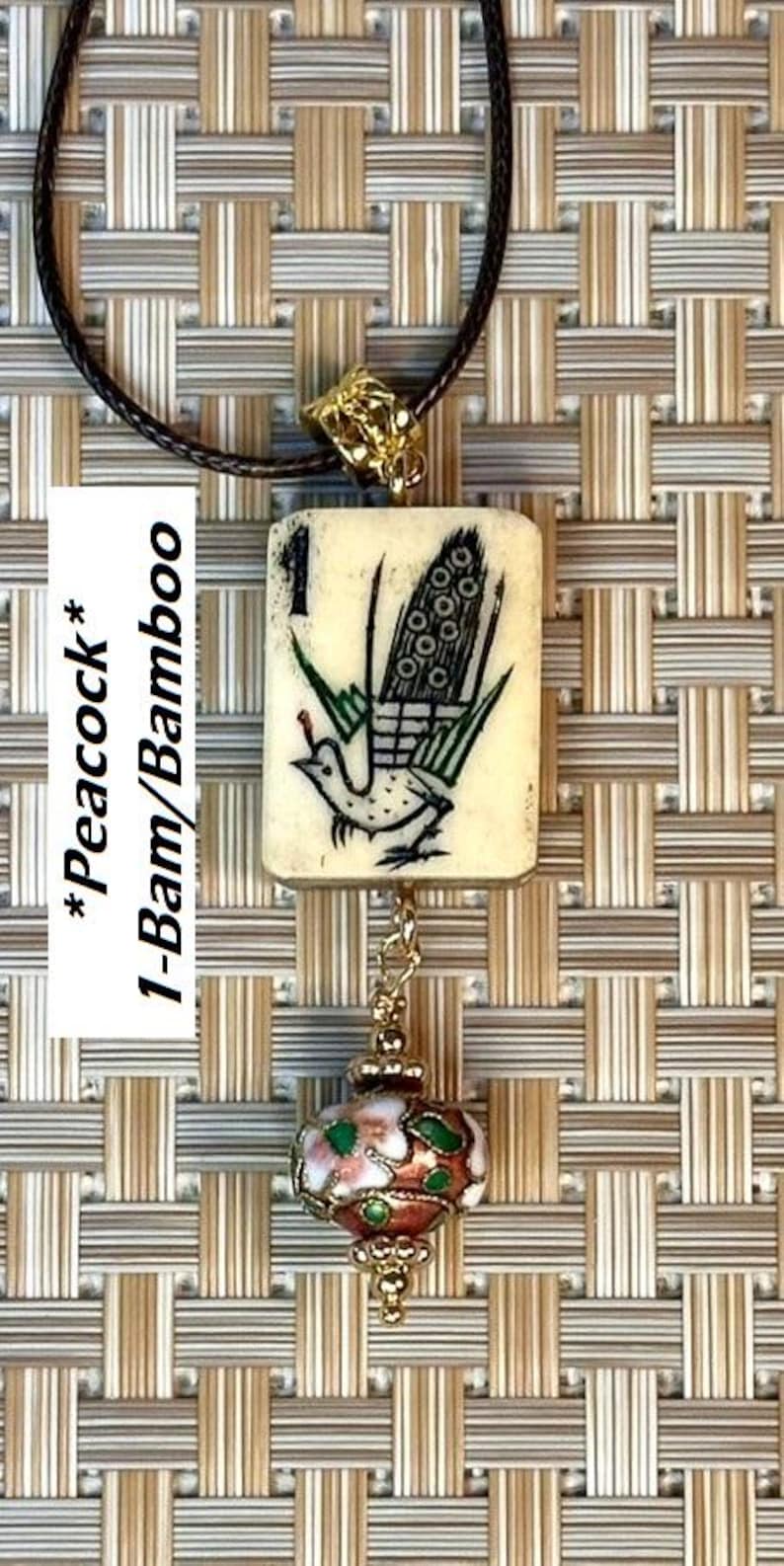 Puede incluir: Un collar con colgante que presenta una baldosa de color crema con una ilustraci&oacute;n de pavo real y el texto "1-Bam/Bamboo". El colgante est&aacute; conectado a una cuenta decorativa esmaltada y una cadena de metal dorado. El collar cuelga de un cord&oacute;n negro.