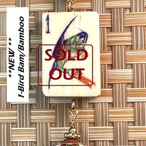 Puede incluir: Un collar con colgante que presenta una baldosa rectangular con una ilustraci&oacute;n de un p&aacute;jaro y el texto "SOLD OUT". El collar tiene un cord&oacute;n marr&oacute;n, una cuenta dorada y un dije rojo, verde y dorado. La baldosa mide aproximadamente 2,5 cm x 3,5 cm.