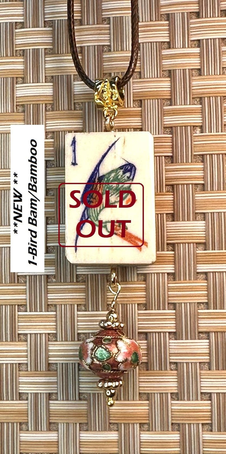 Puede incluir: Un collar con colgante que presenta una baldosa rectangular con una ilustraci&oacute;n de un p&aacute;jaro y el texto "SOLD OUT". El collar tiene un cord&oacute;n marr&oacute;n, una cuenta dorada y un dije rojo, verde y dorado. La baldosa mide aproximadamente 2,5 cm x 3,5 cm.