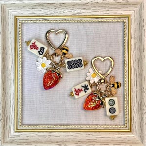 May include: Two heart-shaped gold-tone keychains featuring charms of red strawberries, white daisies, yellow and black bees, and off-white mahjong tiles with black and red characters and symbols. The charms are attached to heart-shaped clasps.