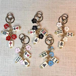 May include: Five keychains with gold-colored clasps, each adorned with miniature Mahjong tiles, faux pearls, and colorful flower charms. The tiles feature Chinese characters and dot patterns, with red, black, pink, and blue floral accents.
