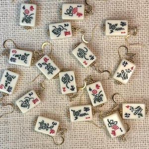 Puede incluir: Un conjunto de pendientes de baldosas de Mahjong blancas con ganchos dorados. Cada baldosa presenta caracteres chinos negros y rojos y dise&ntilde;os florales. Los pendientes est&aacute;n dispersos sobre una superficie texturizada de color blanquecino.