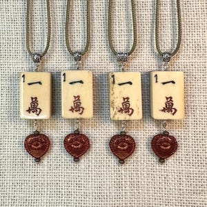 Puede incluir: Cuatro collares con colgantes rectangulares de color marfil con caracteres chinos negros y rojos. Cada colgante está conectado a un dije en forma de corazón rojo. Los collares tienen un cordón verde y detalles plateados.