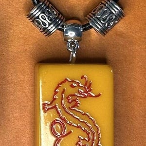 Puede incluir: Collar con colgante con un dije rectangular amarillo con un dise&ntilde;o de drag&oacute;n rojo. El dije cuelga de un cord&oacute;n negro con cuentas plateadas. El texto "Red Dragon" se muestra en la parte inferior.