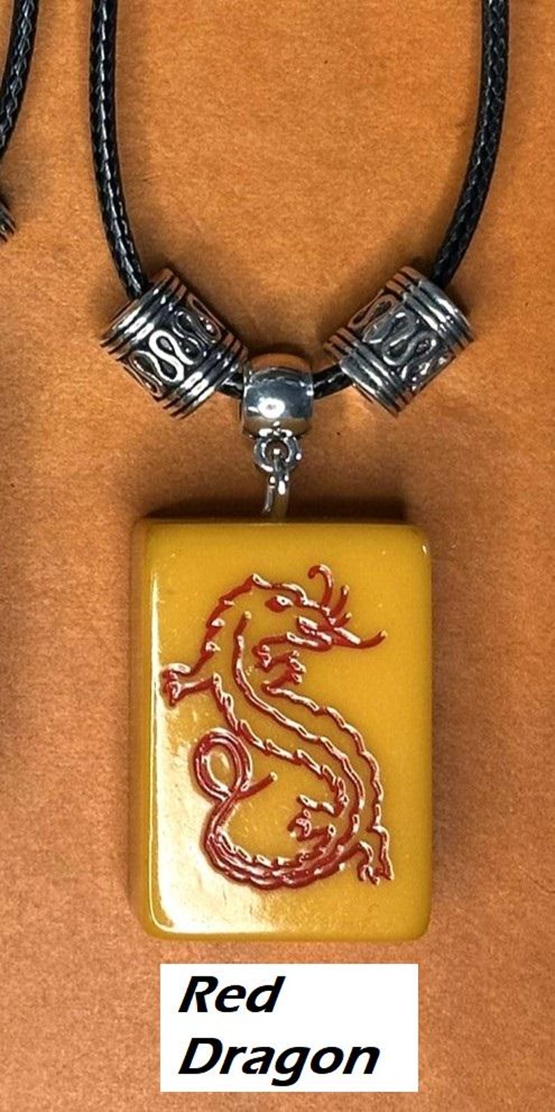 Puede incluir: Collar con colgante con un dije rectangular amarillo con un dise&ntilde;o de drag&oacute;n rojo. El dije cuelga de un cord&oacute;n negro con cuentas plateadas. El texto "Red Dragon" se muestra en la parte inferior.