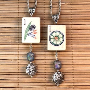 May include: Two silver necklaces with rectangular pendants featuring the number "1" and colorful illustrations. One pendant has a bird design, the other a floral design. Each necklace has a Buddha head bead and a silver flower bead.